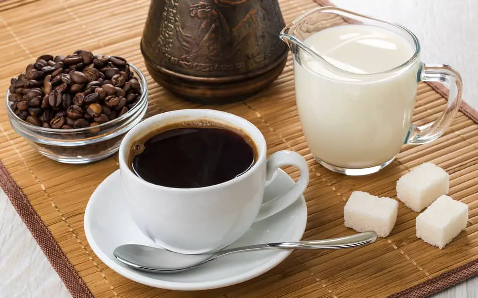 coffee surrounded by sugar cubes, milk and whole coffee beans with coffee maker in background