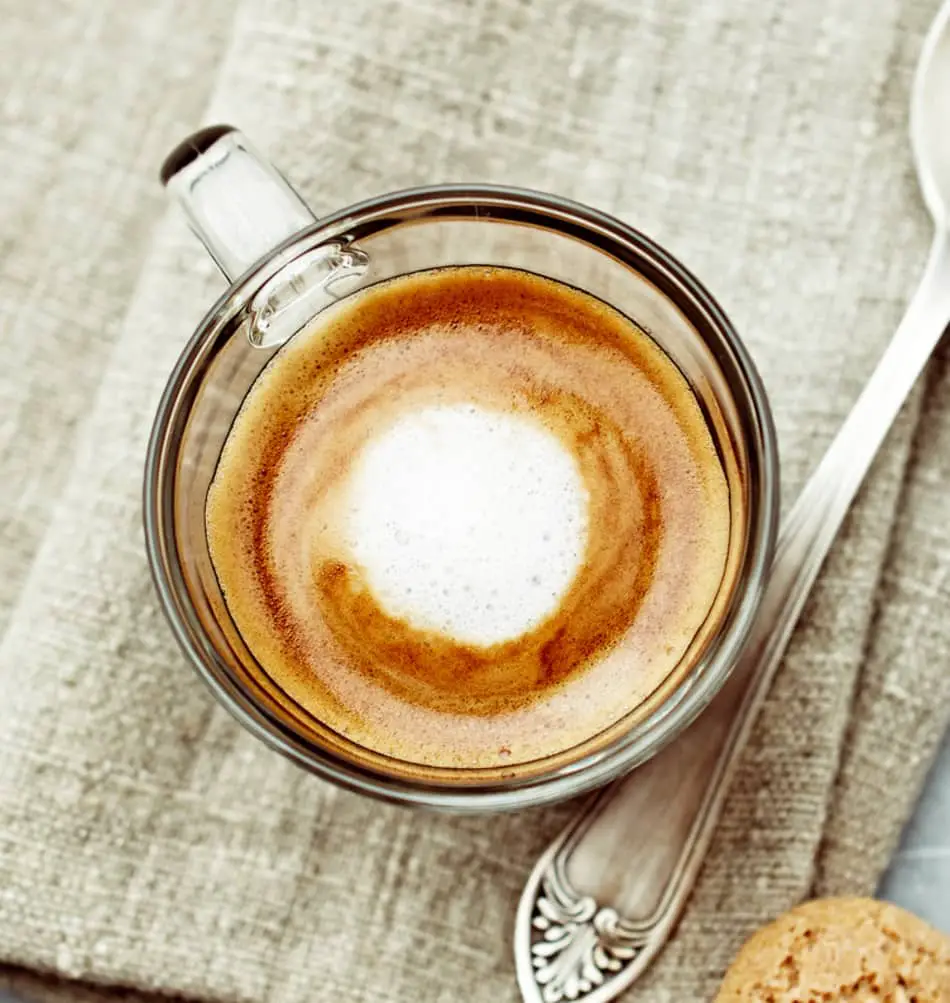 a cup of espresso macchiato on a cloth next to a spoon and sweet biscuit