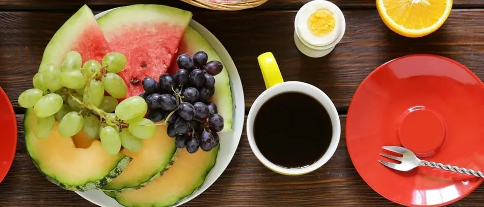 cup of black coffee next to a bowl of fruit and red plate with fork