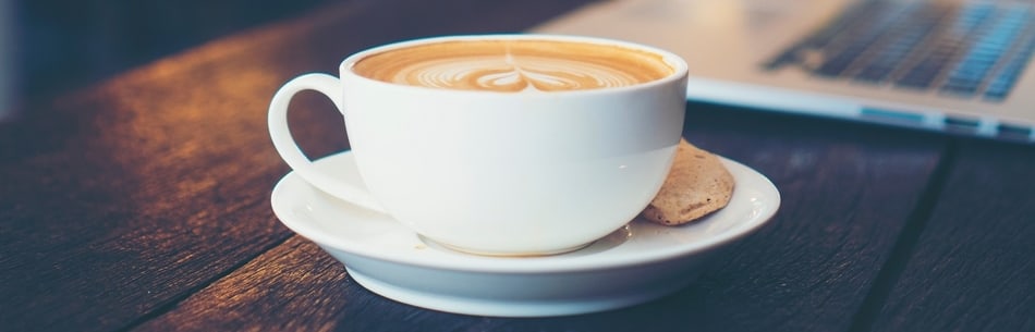 white cup of latte coffee on saucer with biscuit with laptop in background on wooden table